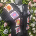 Black crochet cardigan with colorful geometric patterns on a mannequin against a floral background.