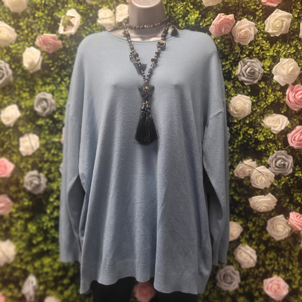 Mannequin wearing a light blue jumper with a long necklace against a floral wall.