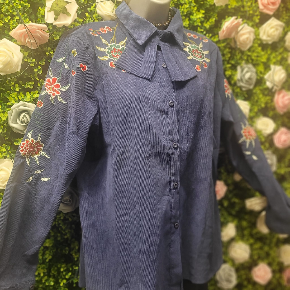Blue corduroy shirt with floral embroidery on a mannequin against a floral background